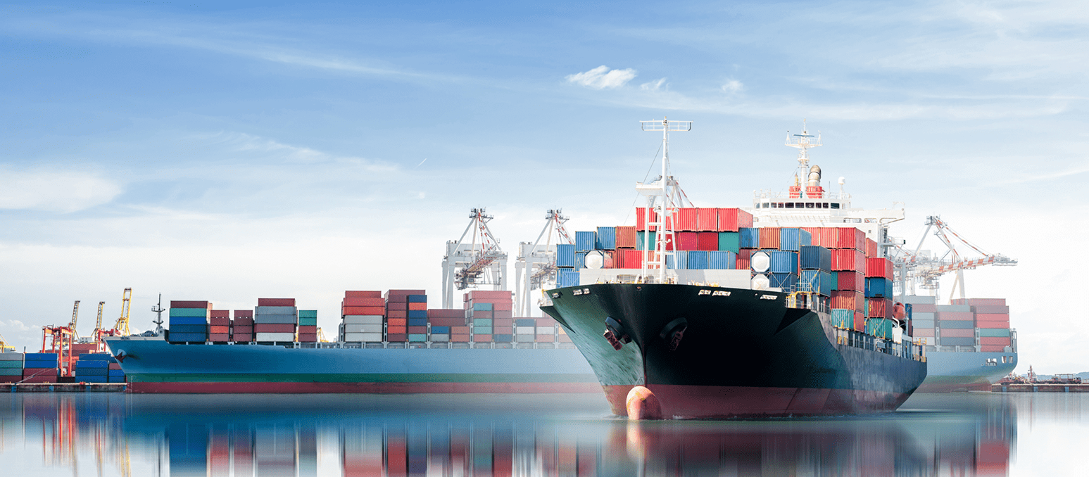 Two large cargo ships docked, loaded with colorful shipping containers under a clear sky.